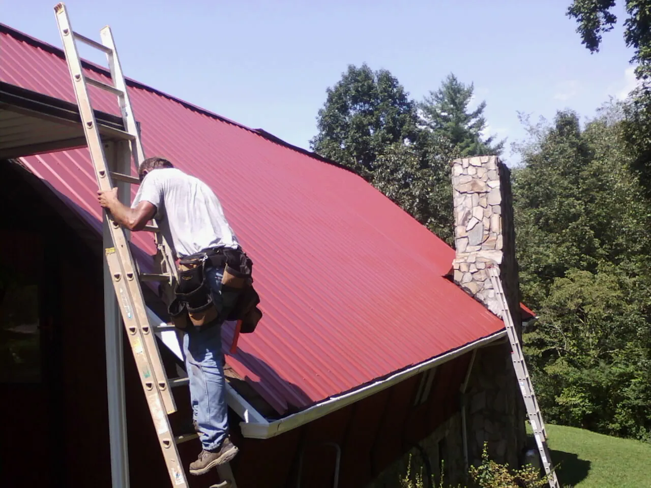 Roofer on a ladder beside a red metal roof and stone chimney — Soffit & Fascia Repair by Roofing Brothers in Landis